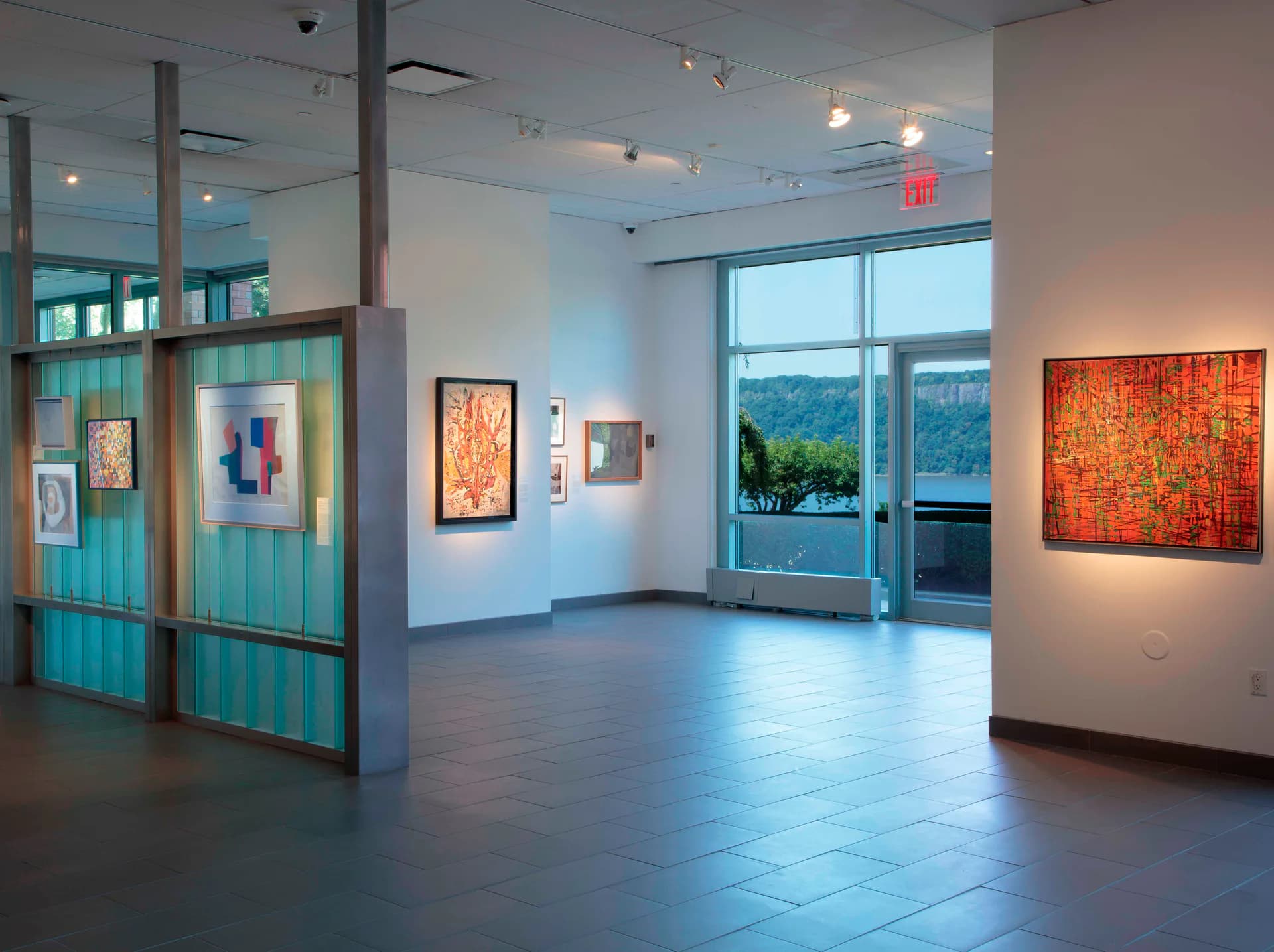 Bright gallery space with turquoise-framed glass dividers, framed artworks on white walls, large windows with landscape view, and red abstract canvas on right wall.