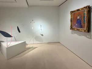 Minimalist gallery interior with white walls, sculptural mobile hanging from ceiling, framed portrait on right wall, and pedestal display.