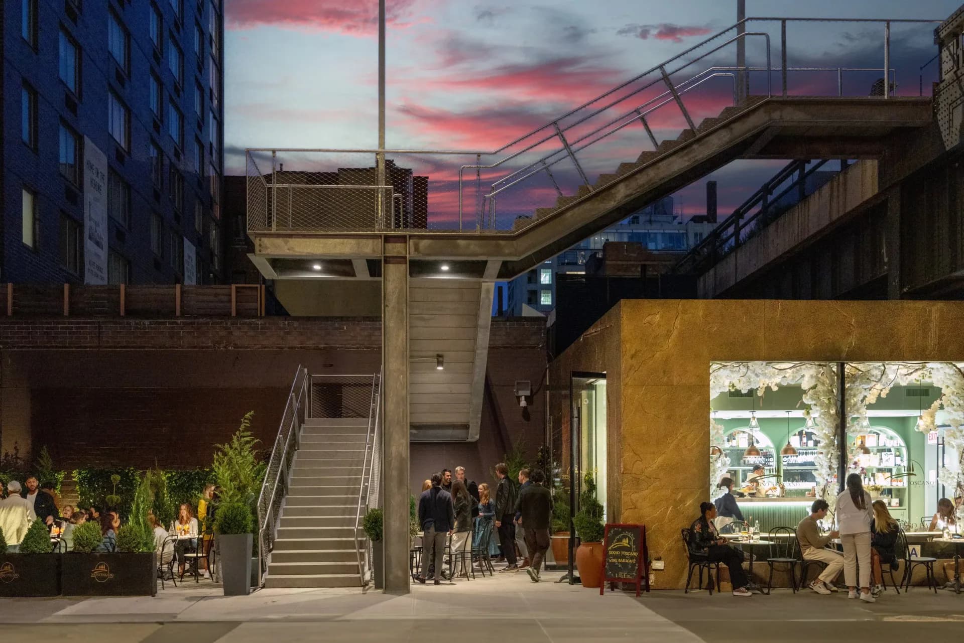 Nighttime urban gallery or retail space with illuminated yellow storefront beneath elevated infrastructure, dramatic sunset sky, planted landscaping, and visitor activity.