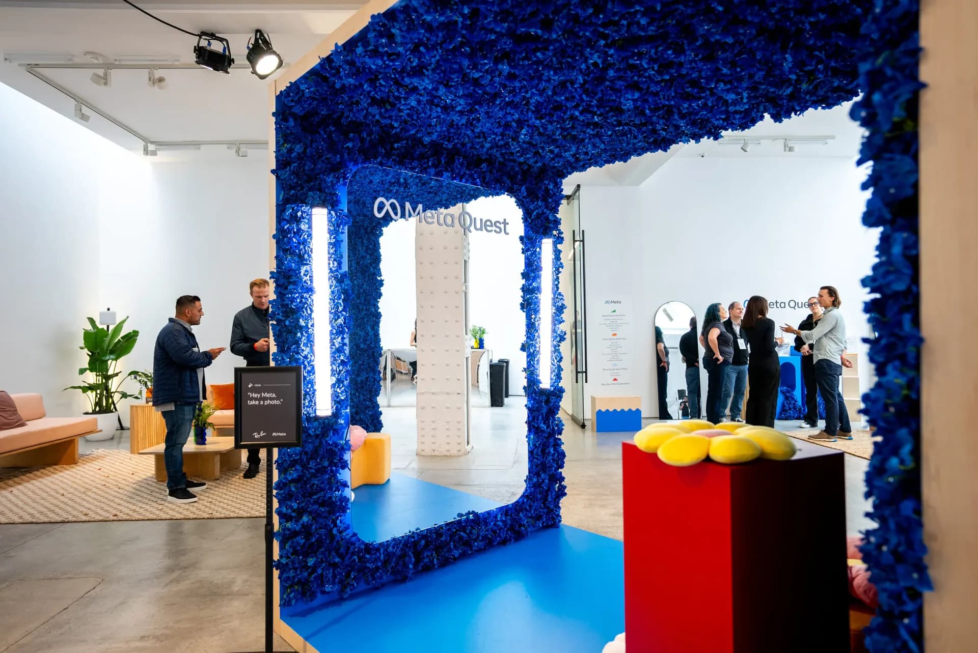 Modern gallery interior with striking blue textured wall installation framing a white corridor, geometric red and yellow sculptural elements, visitors viewing artworks.