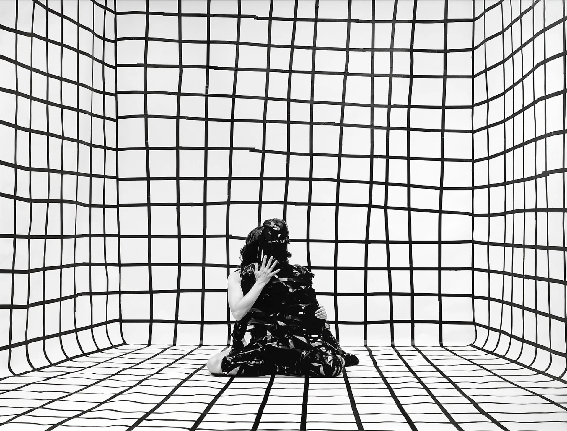 Black and white photograph of a figure sitting in a geometric grid-patterned room with warped perspective lines.