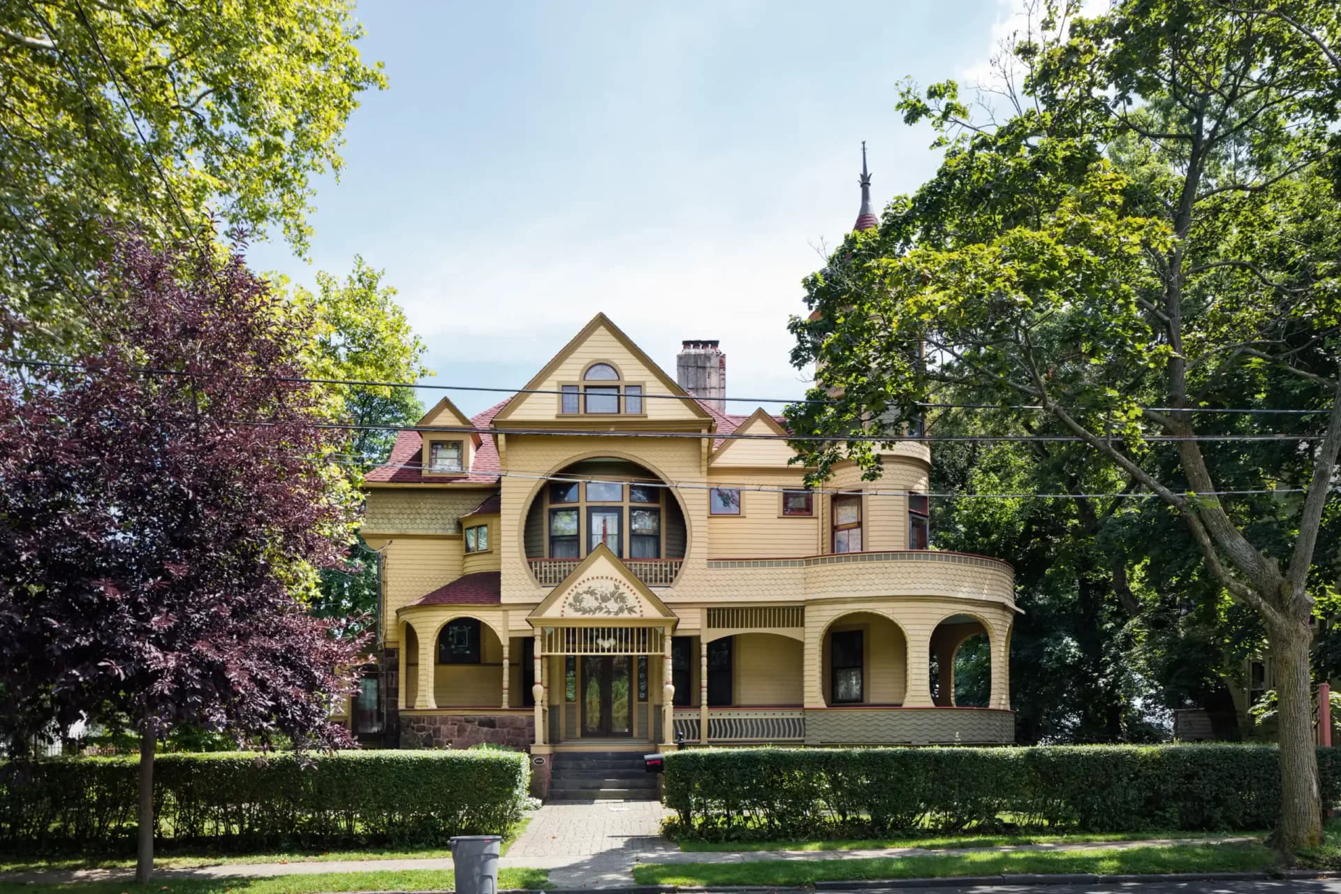 Ornate yellow Victorian mansion with wraparound porch, turret, and gables, surrounded by trees, featured in a Historic Districts Council gallery.