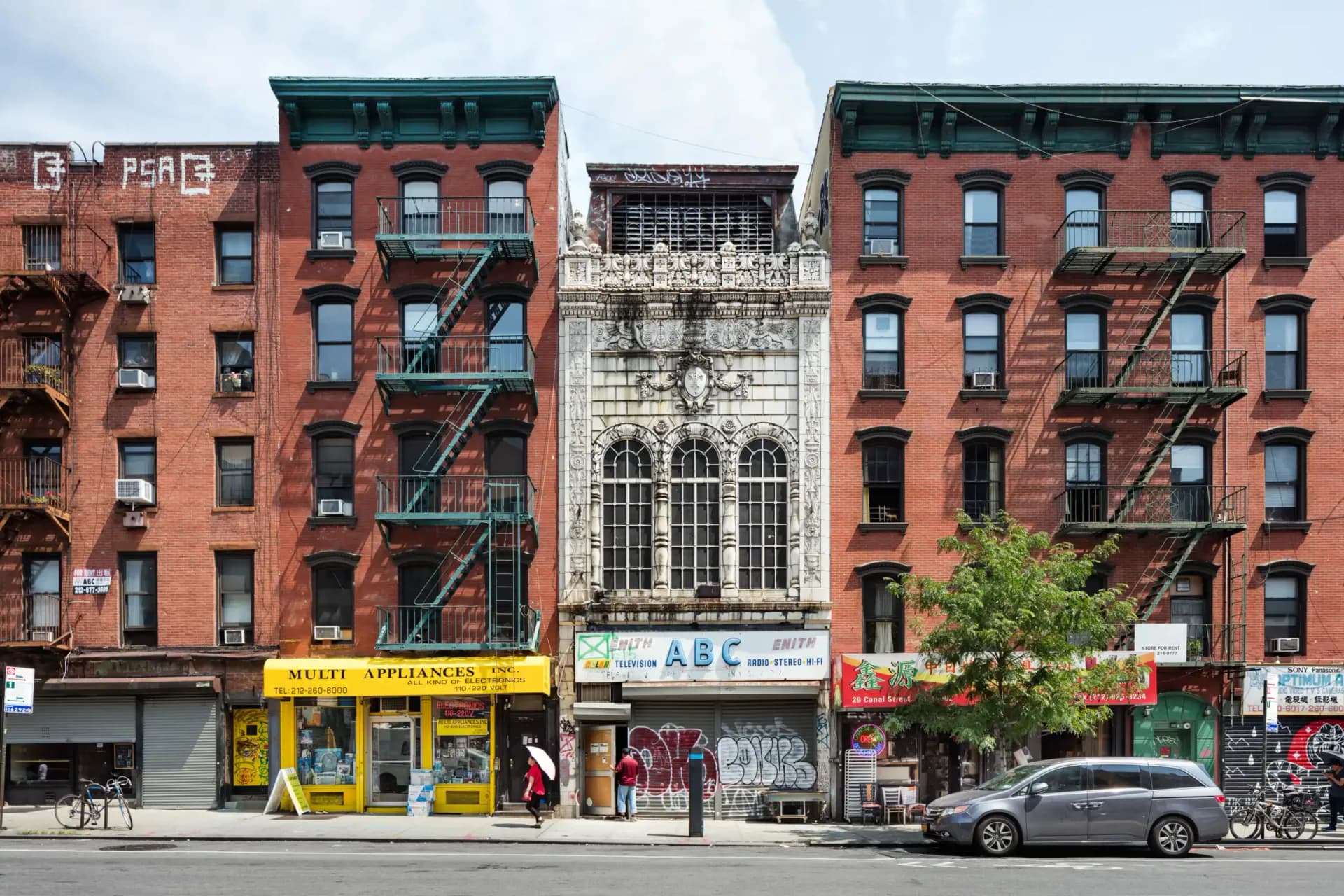 Row of red brick tenement buildings with fire escapes and ground-floor shops on a New York street, part of a Historic Districts Council gallery.