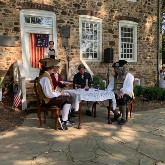 Stone exterior of historic house with white-trimmed windows and doors; costumed interpreters seated at a table conducting a historical reenactment outdoors.