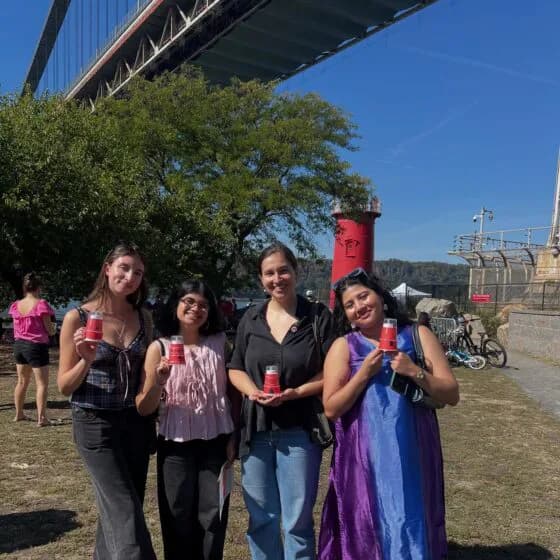 Four women standing outdoors holding pink items near a red cylindrical structure and bridge, with grass and urban waterfront visible.