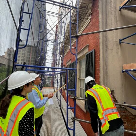 Two workers in safety vests and hard hats on metal scaffolding attached to a historic brick building facade during renovation work.
