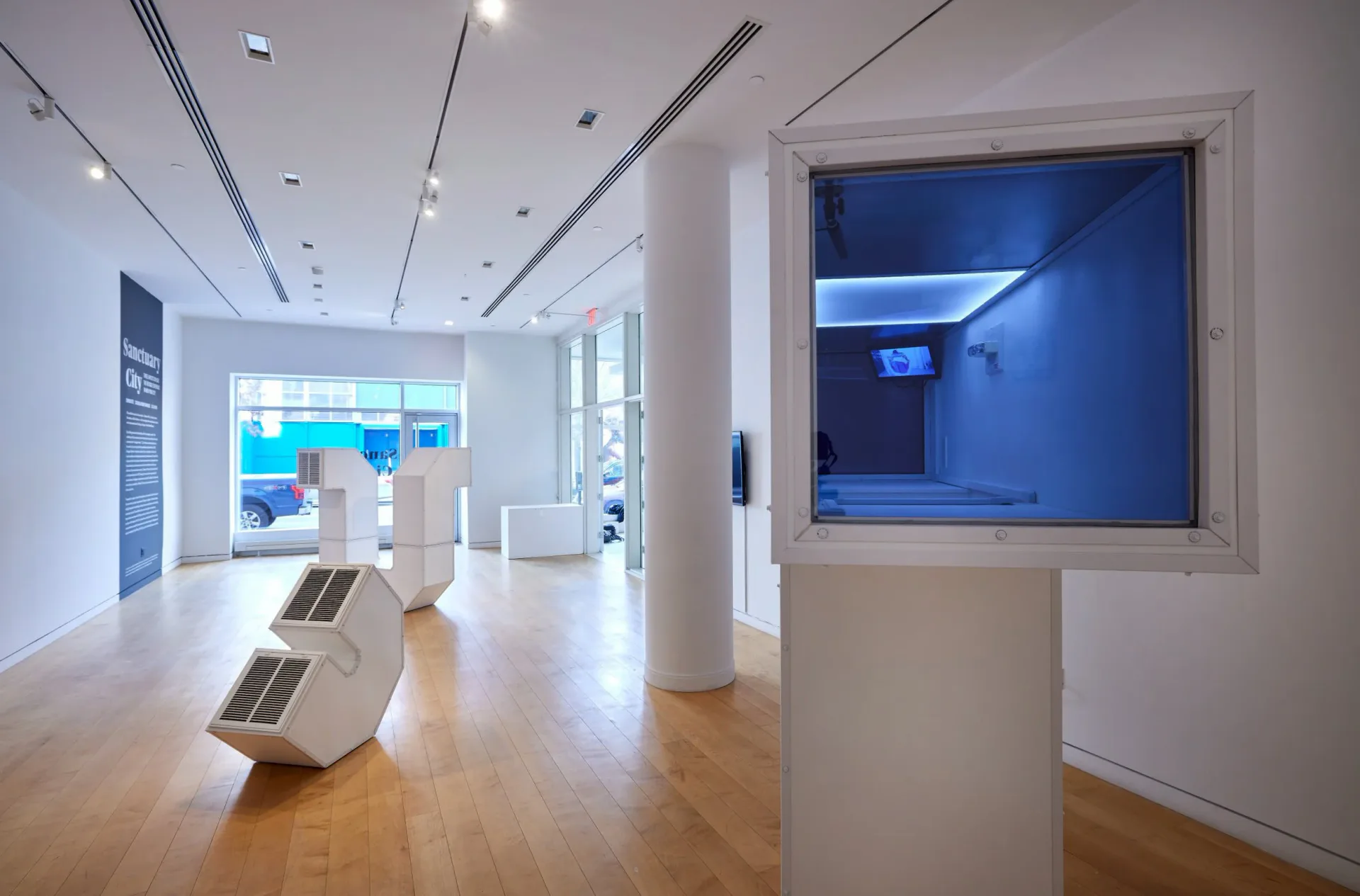 Modern, minimalist gallery interior at Hunter College - Hunter East Harlem Gallery with white walls, wooden floors, blue-lit installation visible.