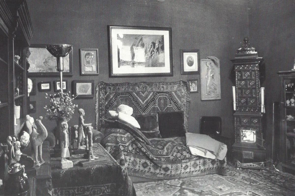 Vintage black-and-white photograph of a cluttered Victorian-era parlor with a patterned daybed, framed pictures on the wall, and decorative sculptures on a shelf.