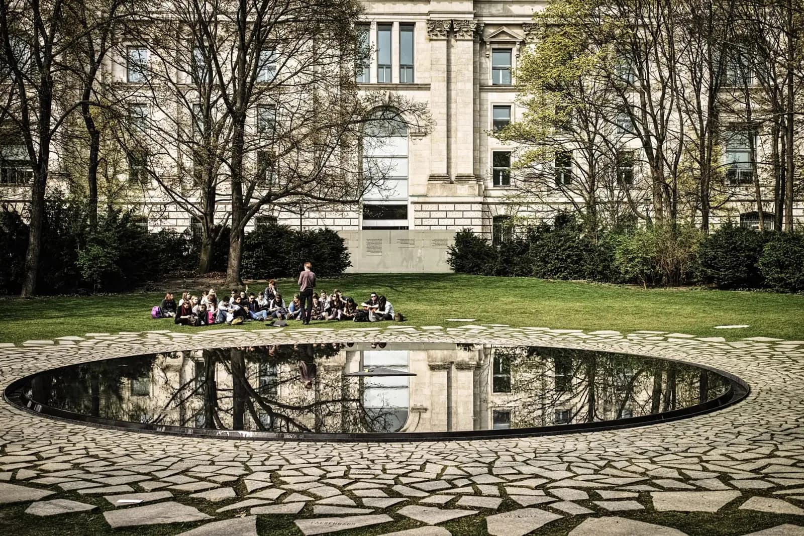 Photograph of a park courtyard with a circular reflecting pool, cobblestone paving, trees, and a grand stone building in the background where people sit on the grass.