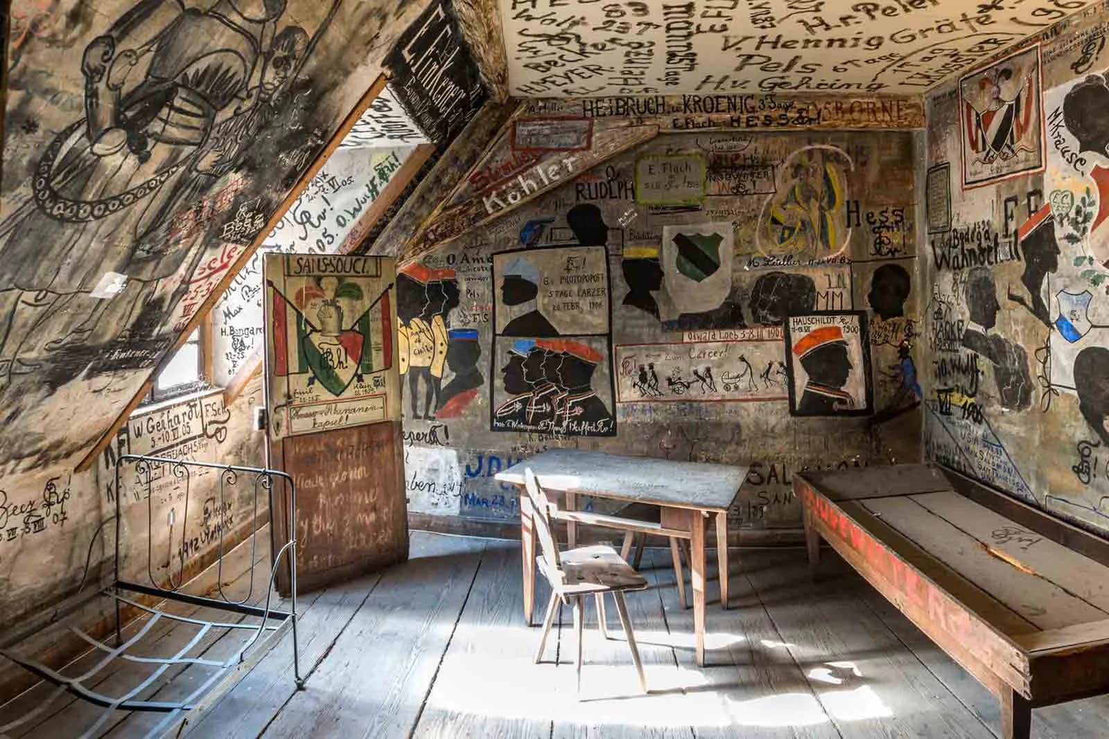 Photograph of a rustic attic room with walls covered in old handwritten graffiti and painted silhouettes, containing a wooden table, chair, bed frame, and bench.