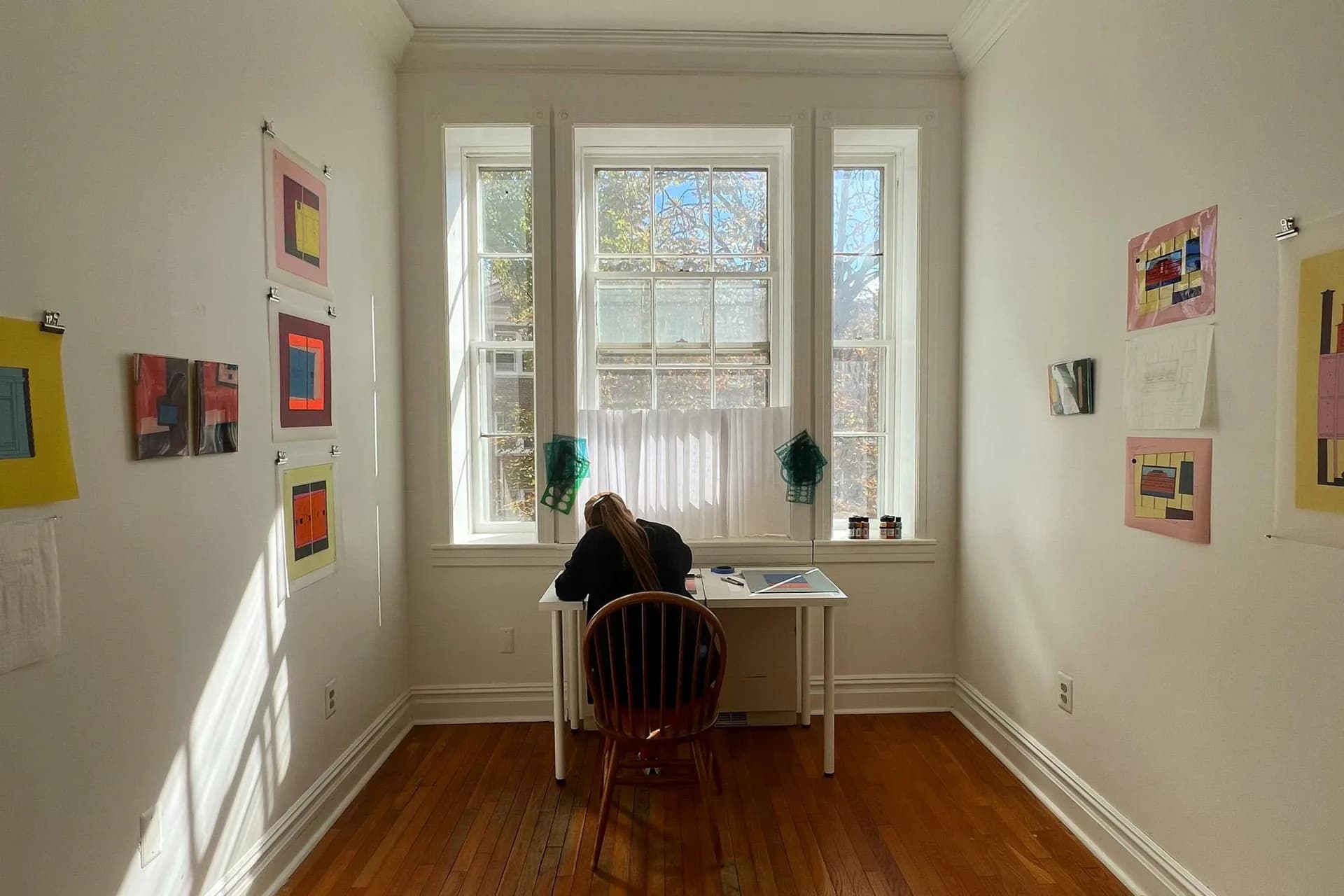 Sunlit gallery room with large windows, wooden floor, and colorful abstract artworks mounted on white walls; single visitor seated at desk.