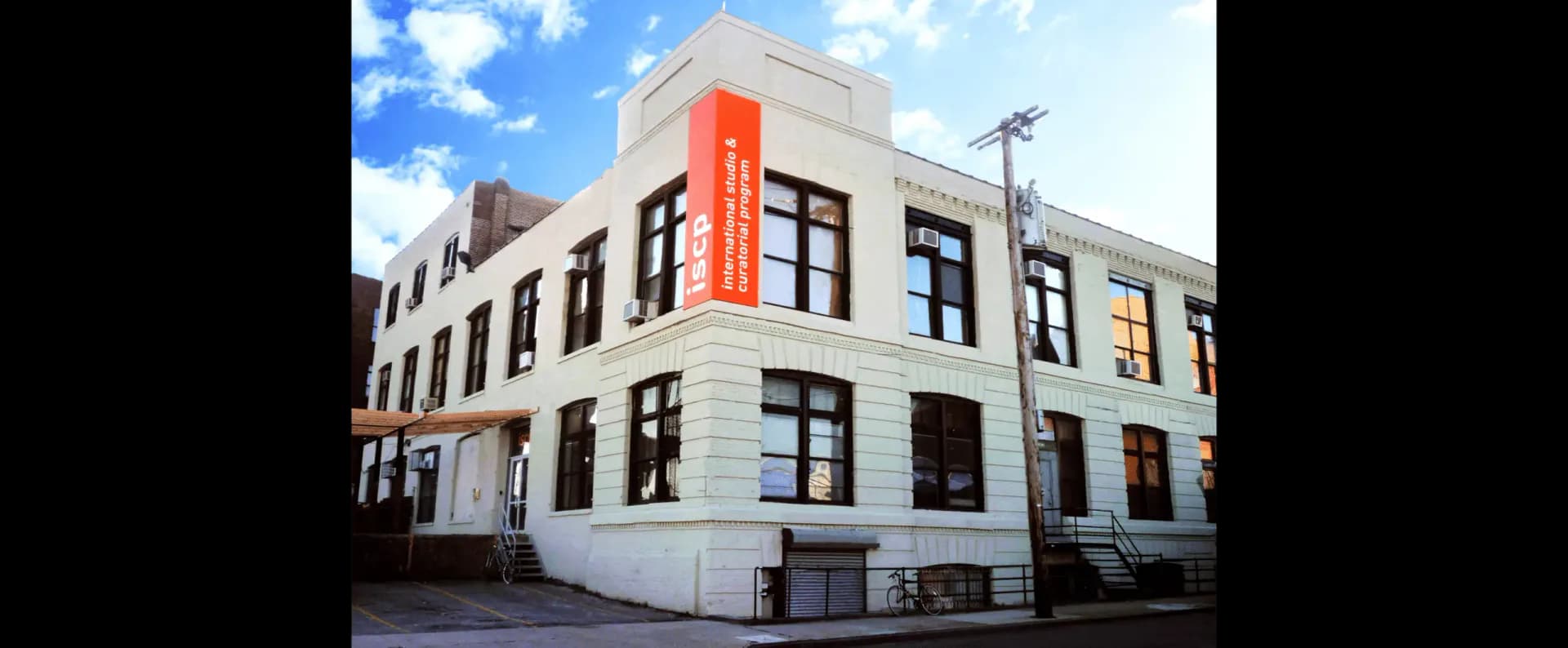 Exterior facade of International Studio & Curatorial Program, a cream-colored corner building with dark-framed windows and an orange vertical sign reading ISCP