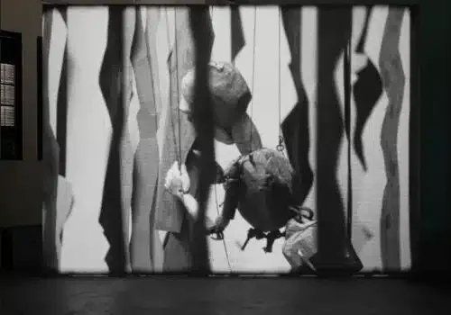 Black and white video still or film projection showing abstracted figures among vertical light streaks or fabric forms, installed at International Studio and Curatorial Program gallery.