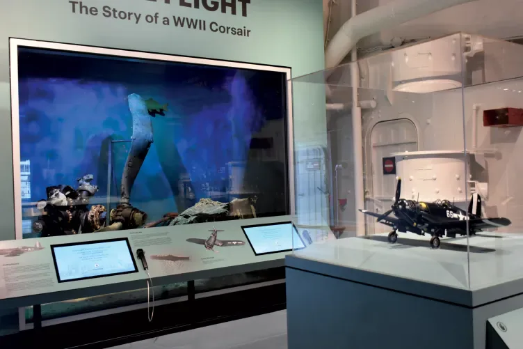 Museum exhibition interior titled 'The Story of a WWII Corsair' with large video screen, interactive displays, and engine components on pedestal.
