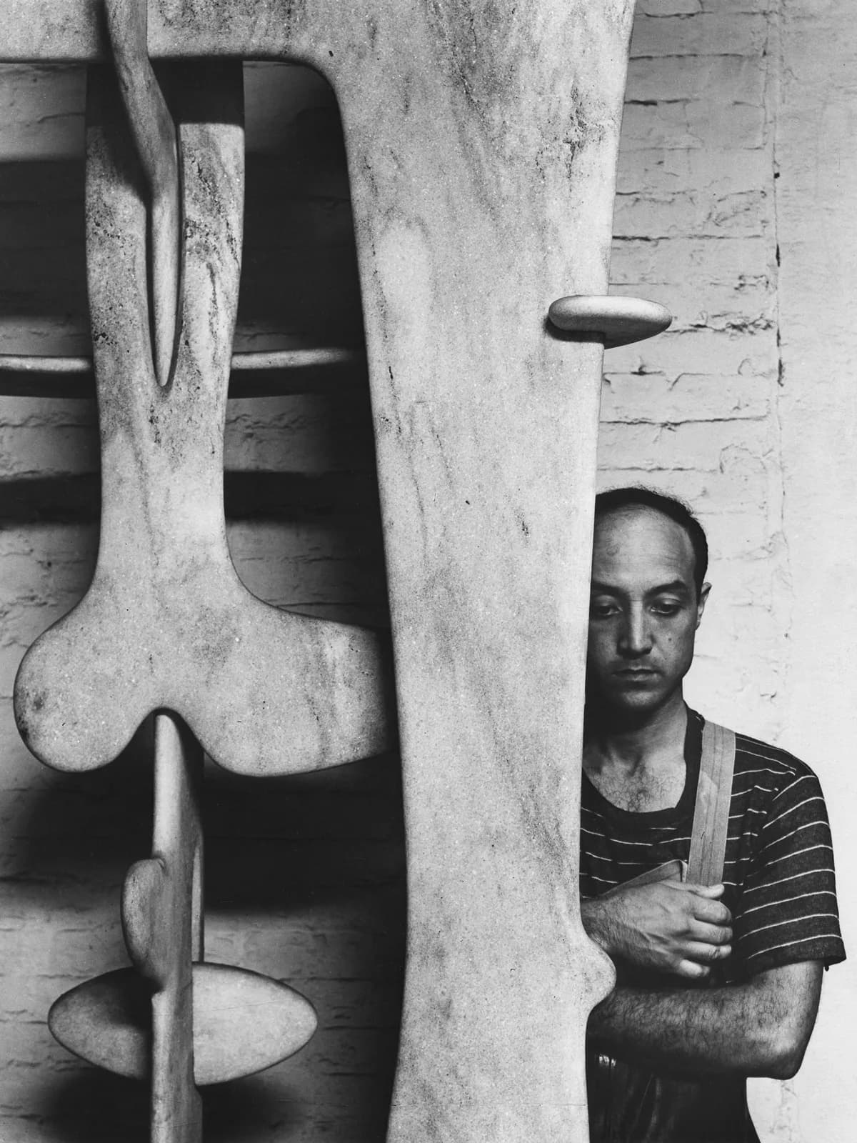 Black and white photograph of a man in striped shirt posing beside a large abstract wooden sculptural form with organic, curved shapes.