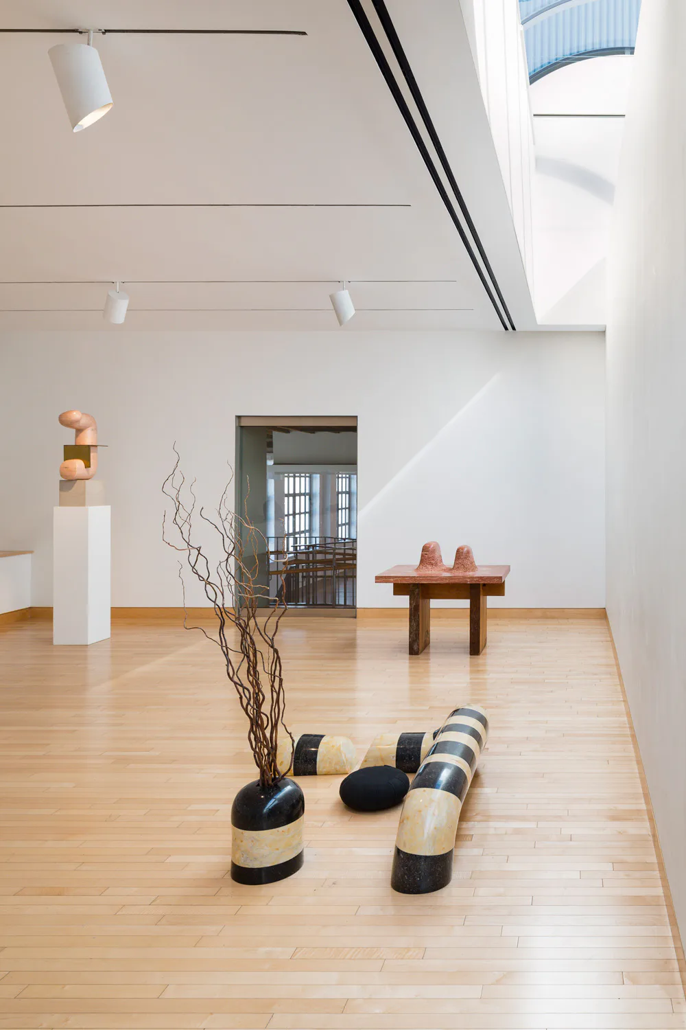 Bright minimalist gallery interior with wooden floor, white walls, skylights, displaying abstract sculptures including cylindrical striped forms and organic objects on pedestals.