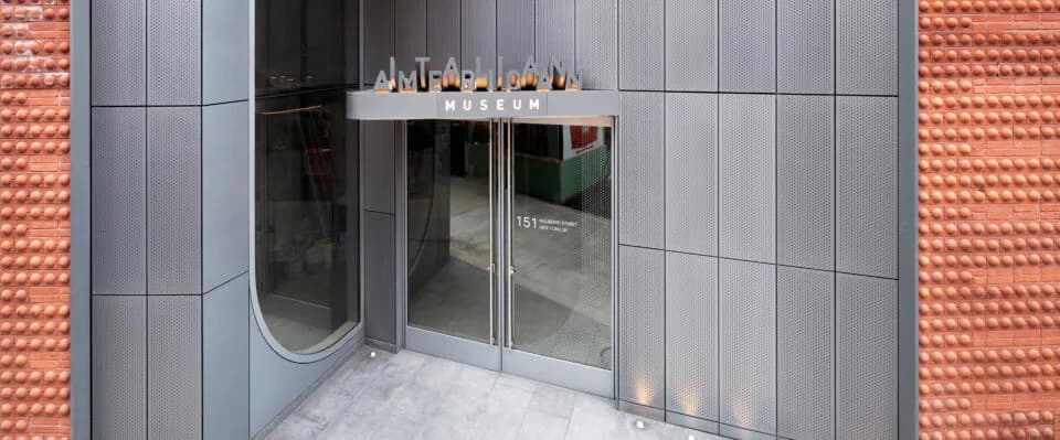 Modern gallery entrance with glass doors and sleek gray tile paneling set against a red brick exterior wall.