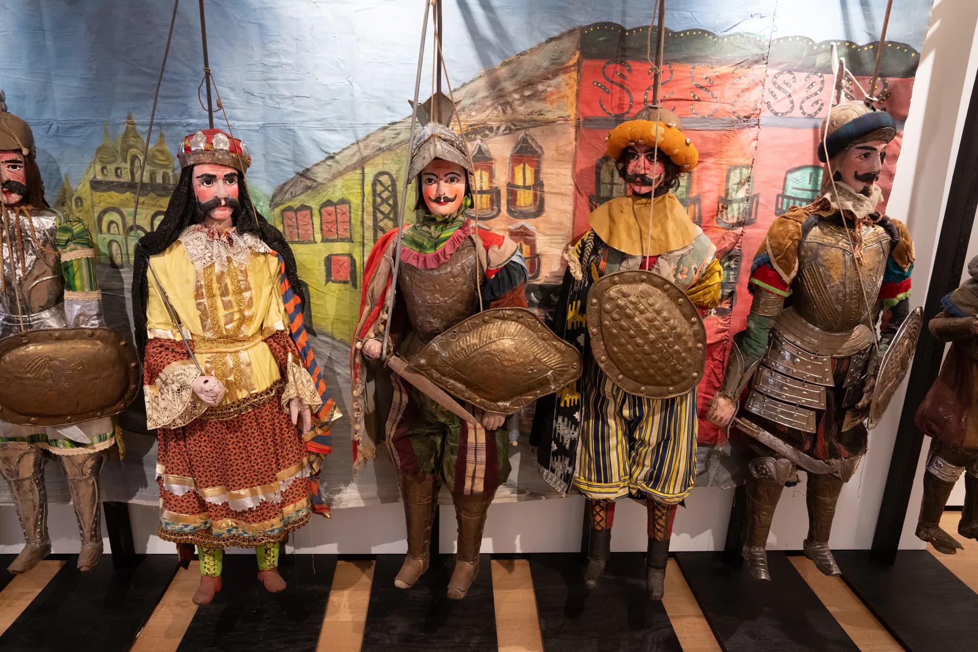 Four life-sized figurative sculptures in historical costume holding spherical objects, displayed against a painted backdrop of colorful buildings.