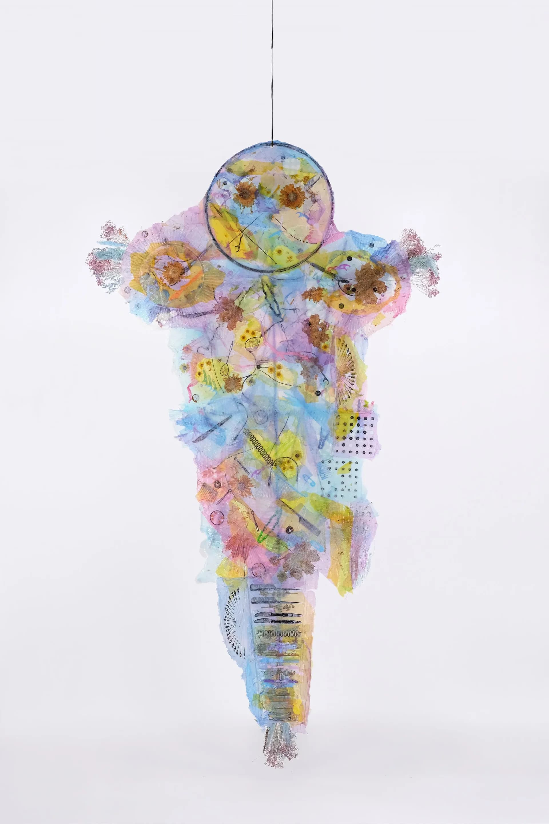 Hanging sculptural mixed-media piece in the shape of a figure with a circular head, composed of pressed flowers, watercolor washes, and translucent materials in pastels.