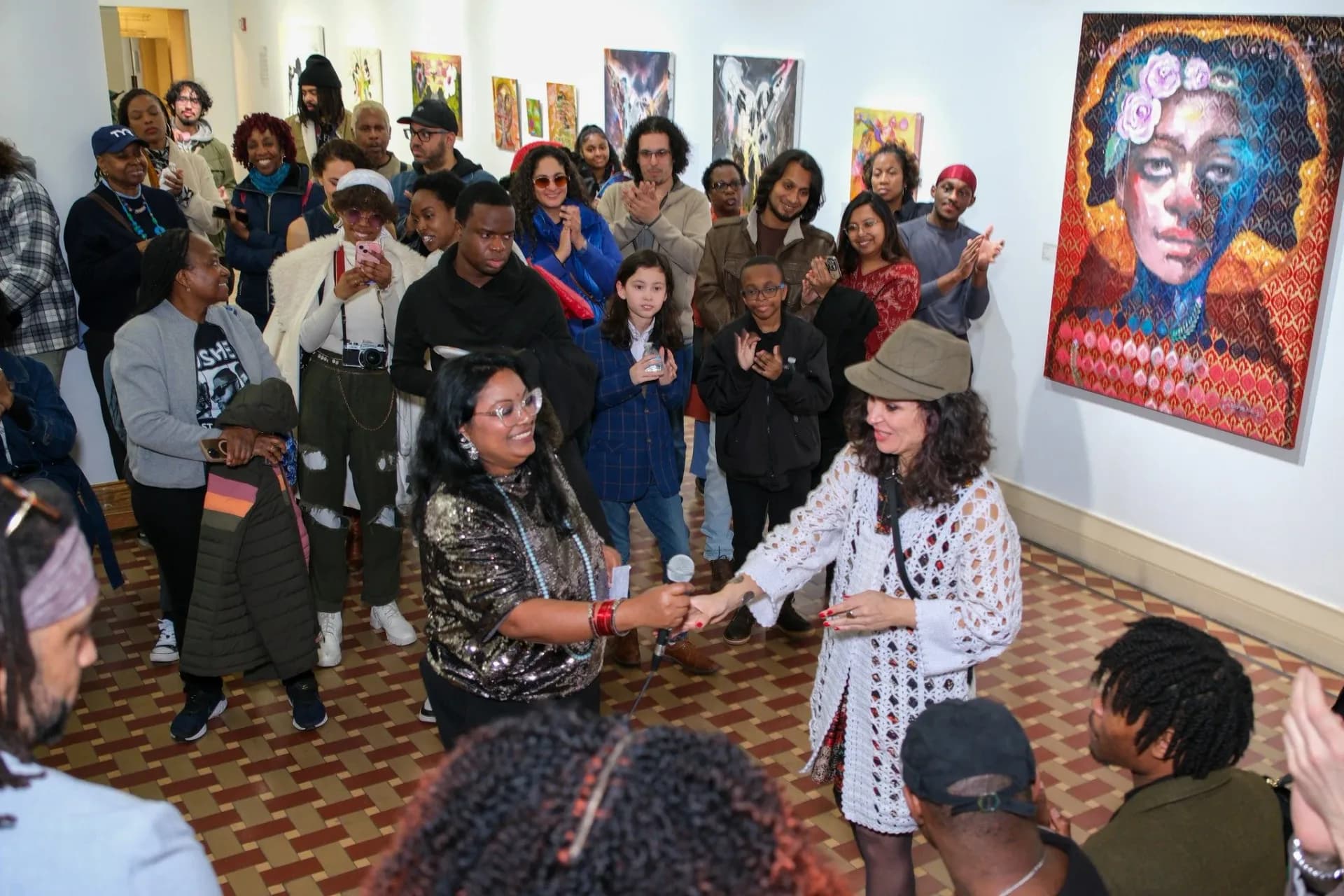 Gallery interior with crowd of visitors gathered around a speaker; colorful portraits displayed on white walls.