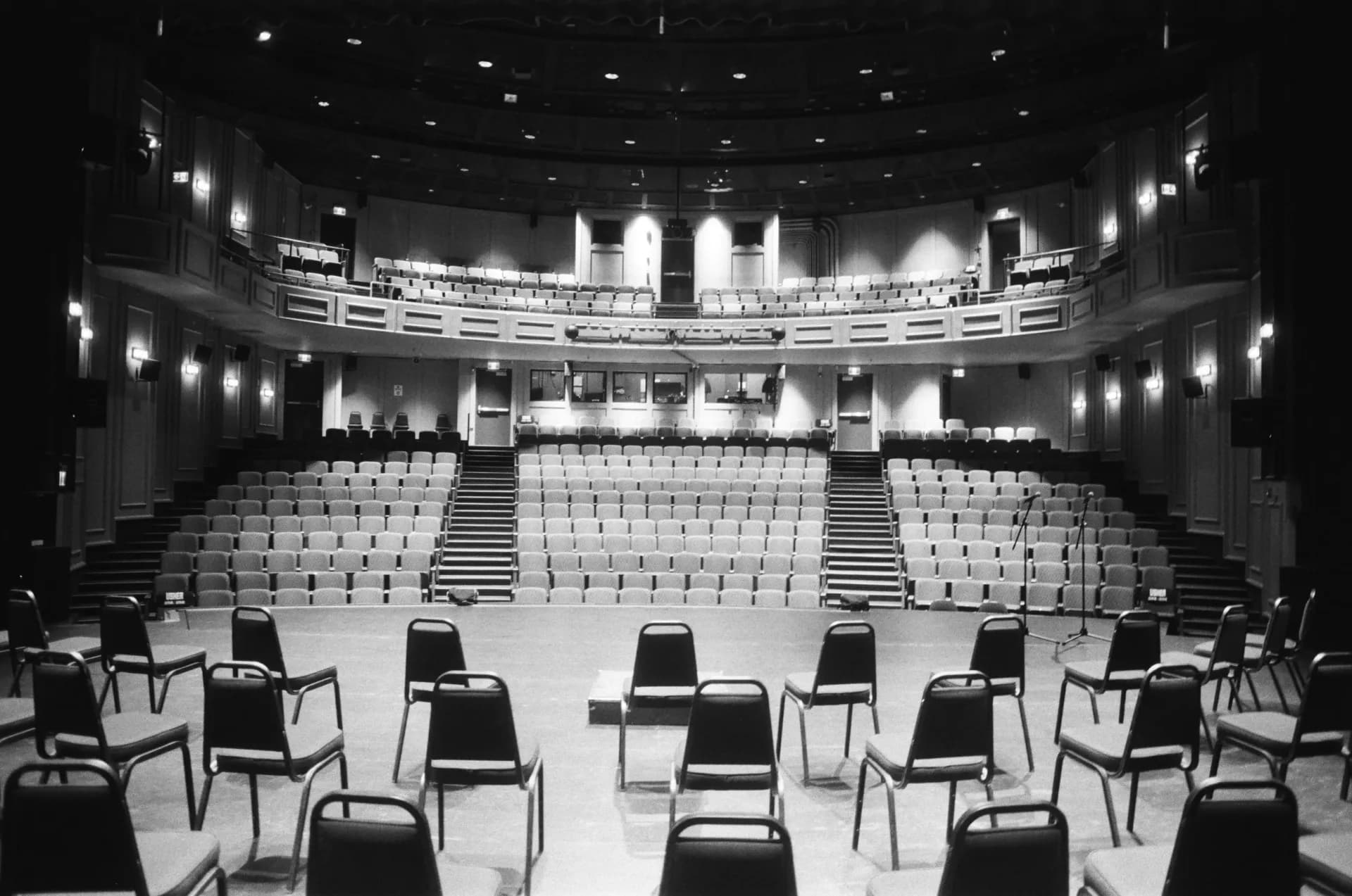 Black and white photograph of an empty auditorium interior with rows of white seats and ornate ceiling with lights.