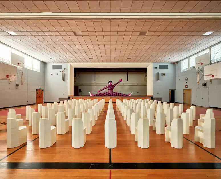 Gymnasium or gymnasium space with wooden floor divided by center line, white cylindrical sculptures arranged in rows, and a performer in red on an elevated stage at the far end.