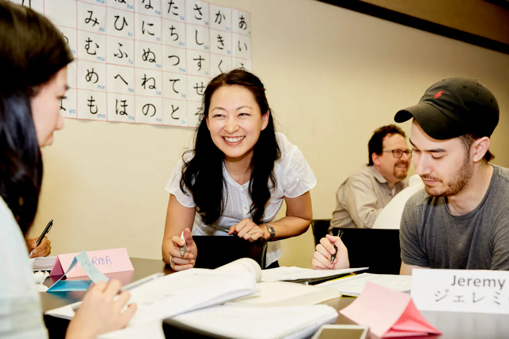 Japanese language class with instructor smiling at center table, interacting with students holding papers and writing materials.