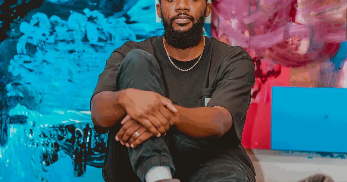 Man wearing black t-shirt and gold chain necklace posing with crossed arms against vibrant blue and pink painted backdrop.