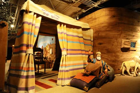 Jewish Children's Museum interior exhibit space featuring striped fabric tent entrance with Hebrew text, wooden furnishings, and visitor exploring the installation.