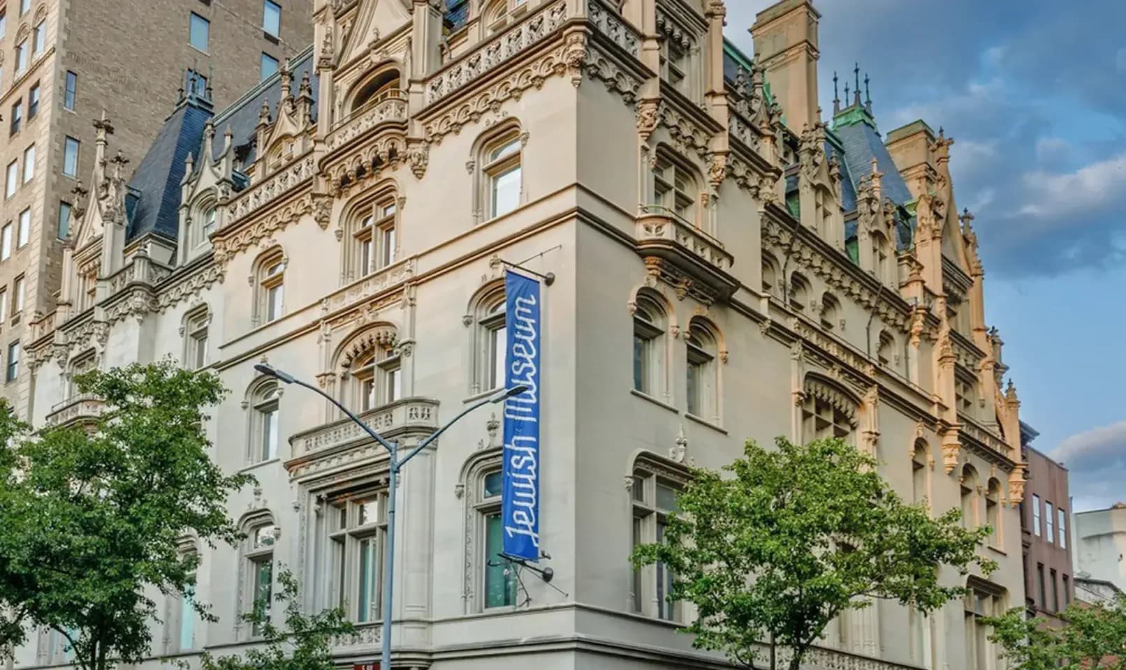 Exterior of a beige stone building with ornate architectural details, arched windows, and a blue banner, featuring green trees in foreground.