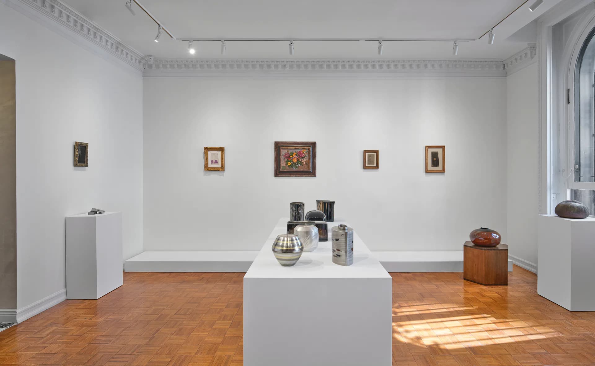 Gallery interior with white walls, wooden herringbone floor, and white pedestals displaying small artworks and decorative objects.