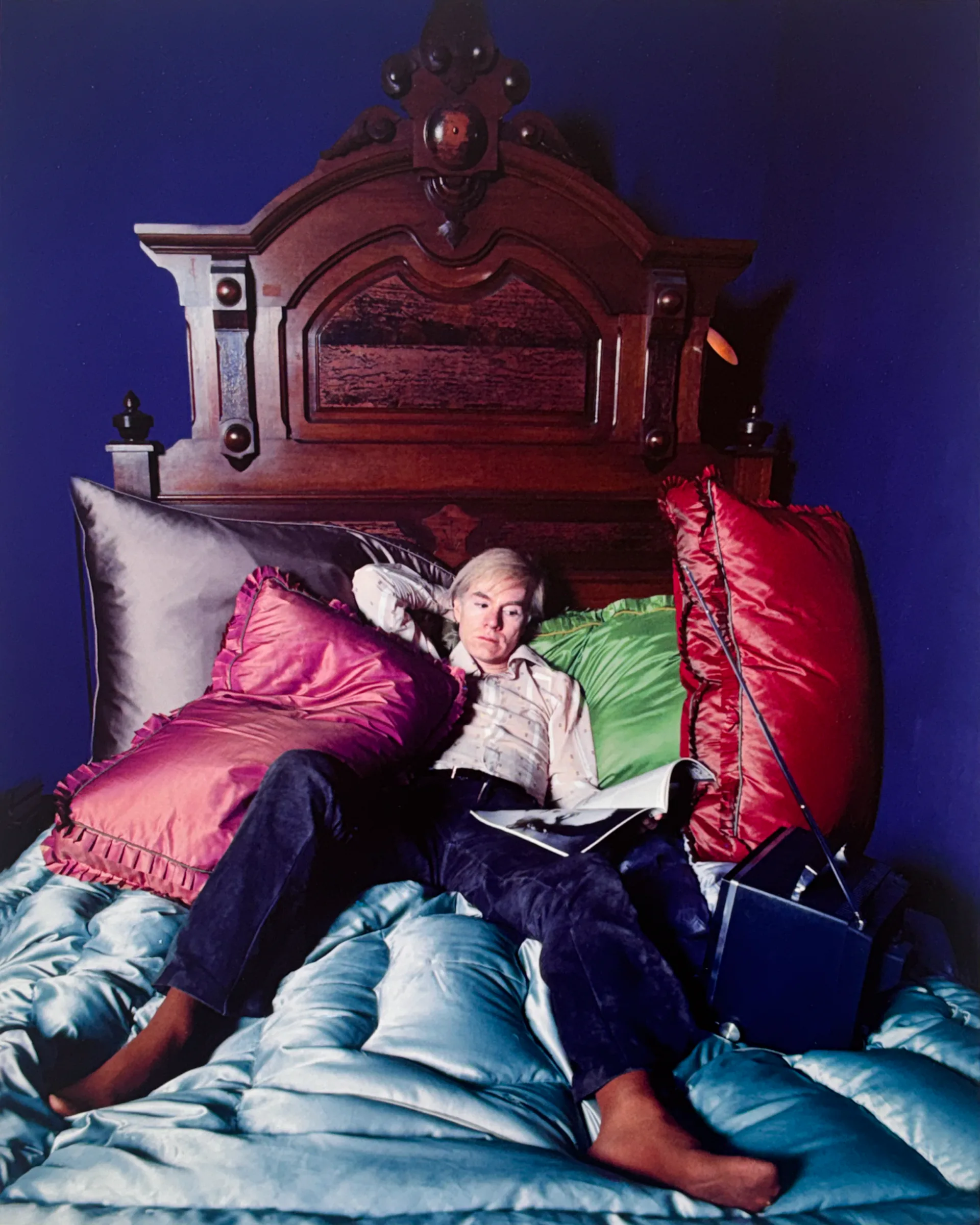 Portrait photograph of a person reading on an ornate dark wooden bed with colorful silk pillows against deep blue wall.