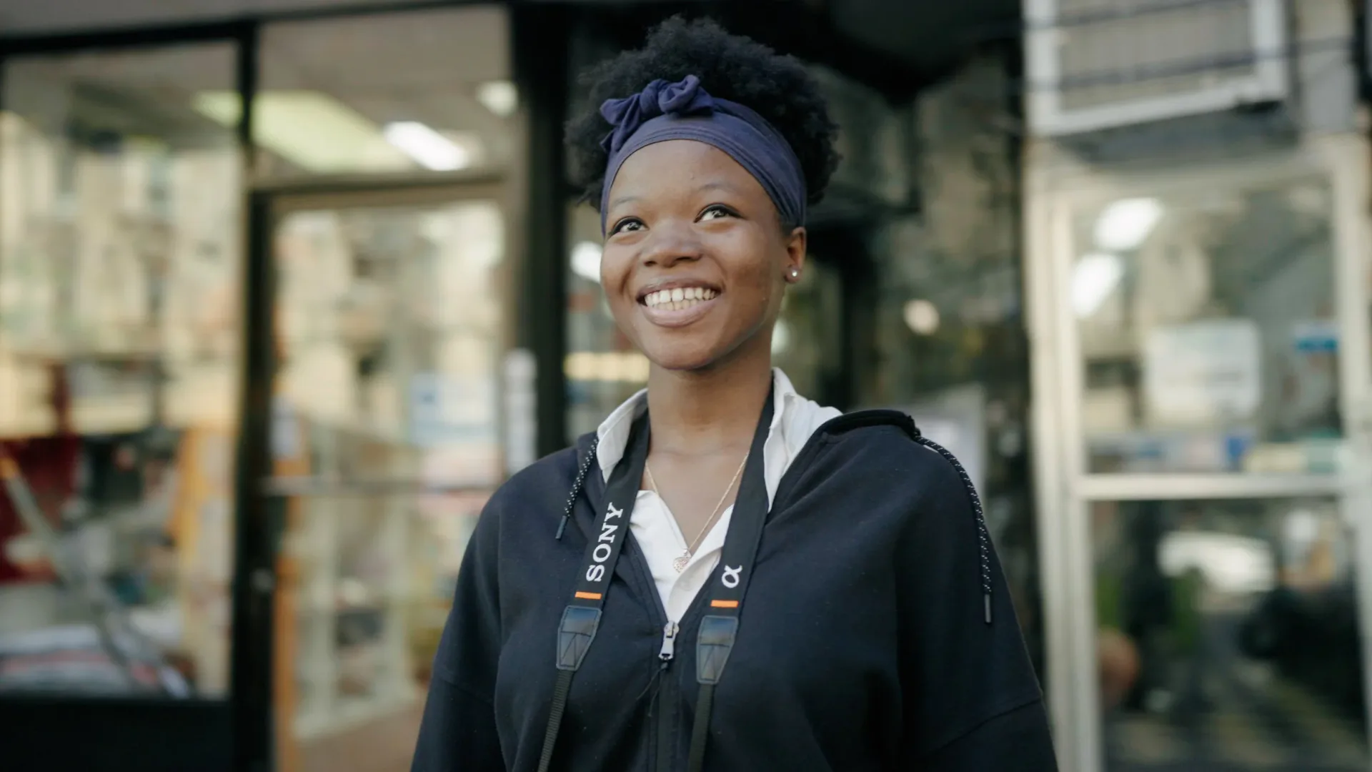 Smiling person wearing a black jacket and blue headwrap, standing outdoors in front of a storefront.