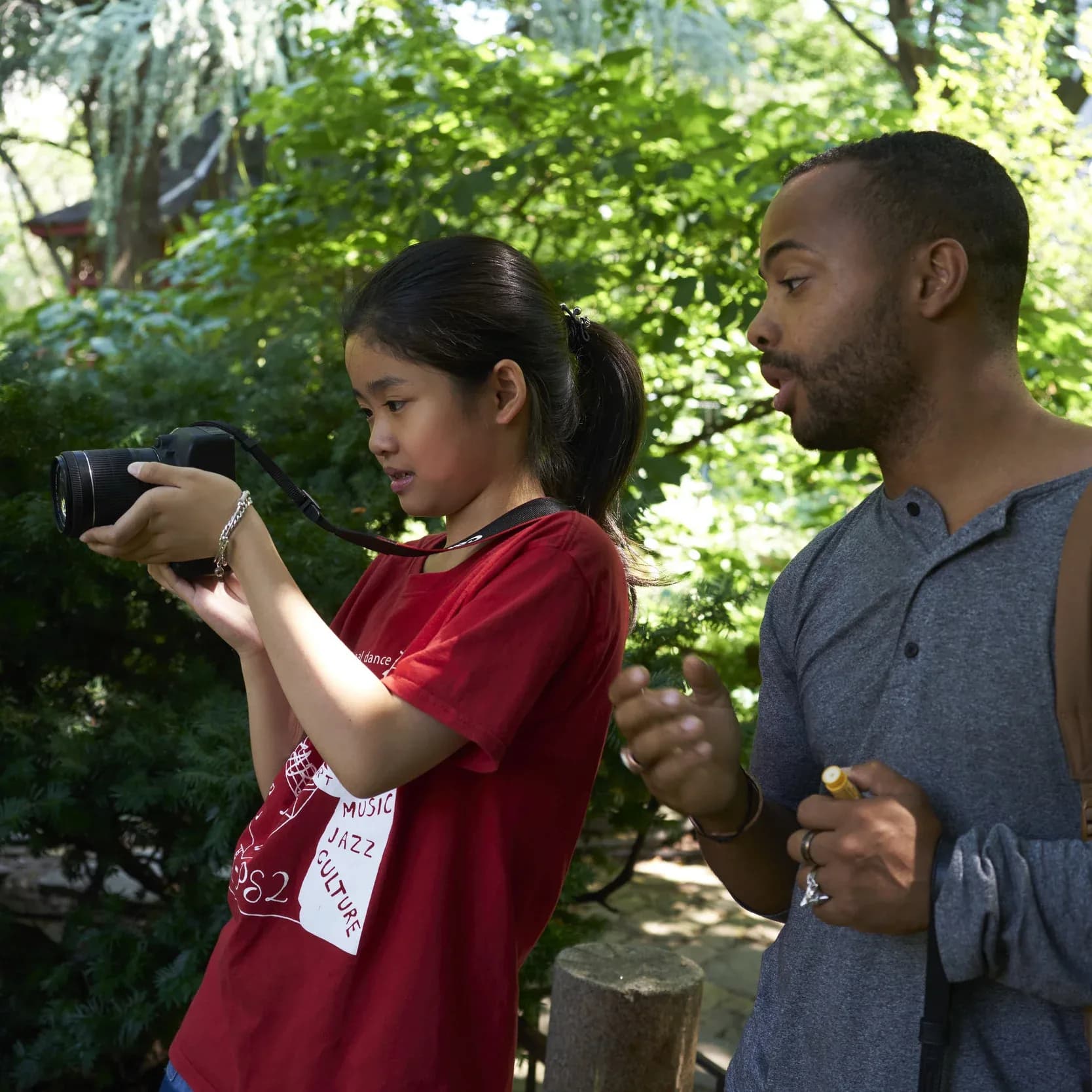 Two people outdoors among green foliage; person in red shirt holds binoculars while person in blue shirt observes.