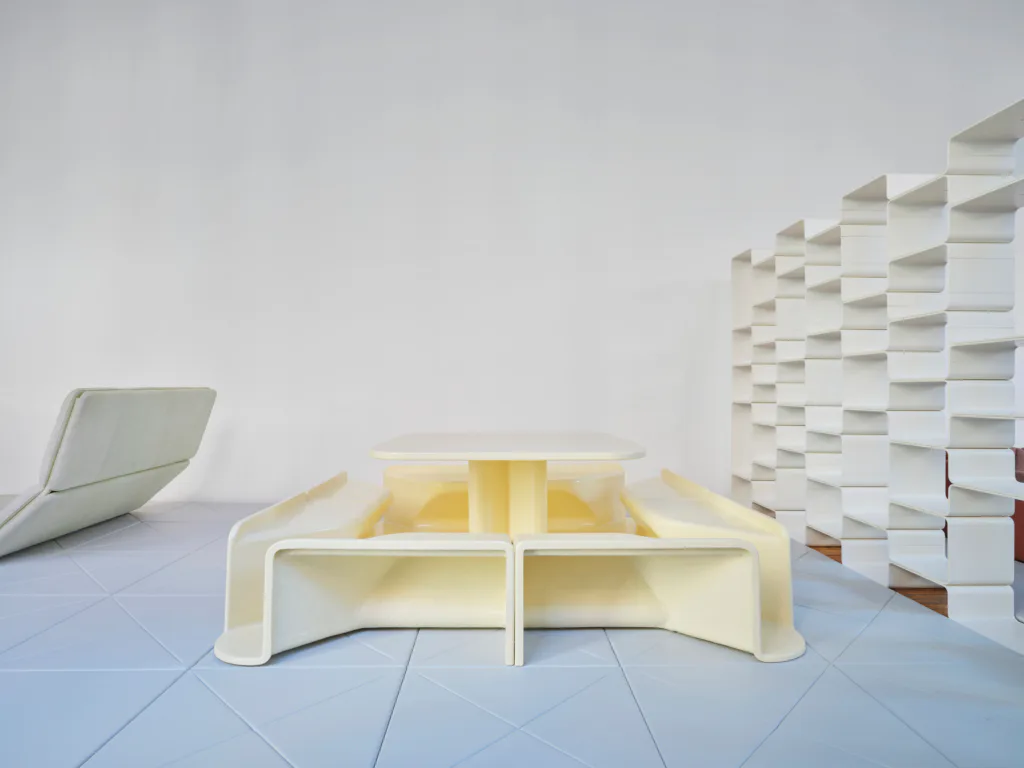 Minimalist sculptural installation featuring white curved form with yellow interior, stacked white elements, and angled panel on wooden floor.