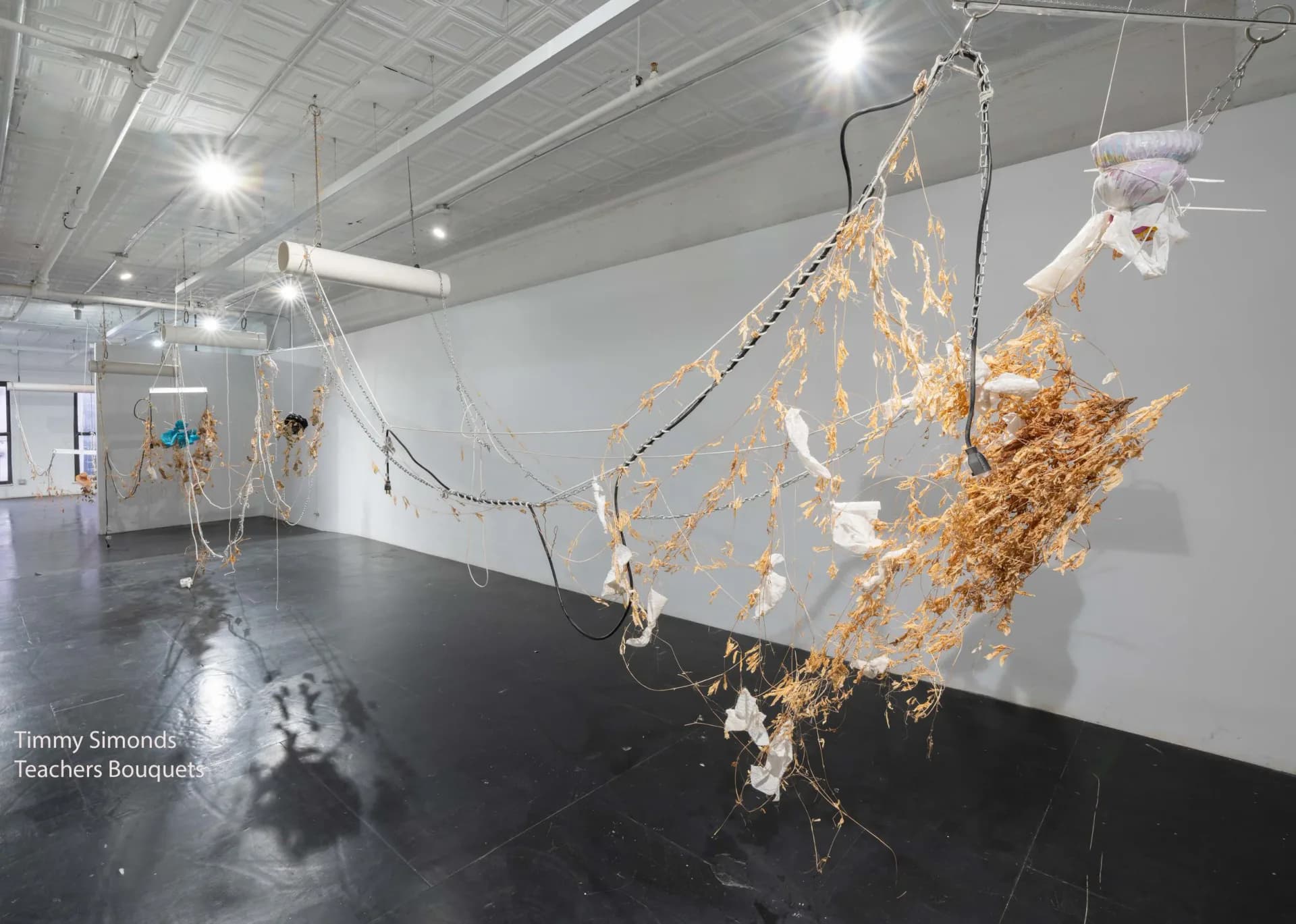 Gallery interior with suspended mixed-media sculptural installation featuring gold leaf and white elements hanging from cables in bright, industrial white-walled space.