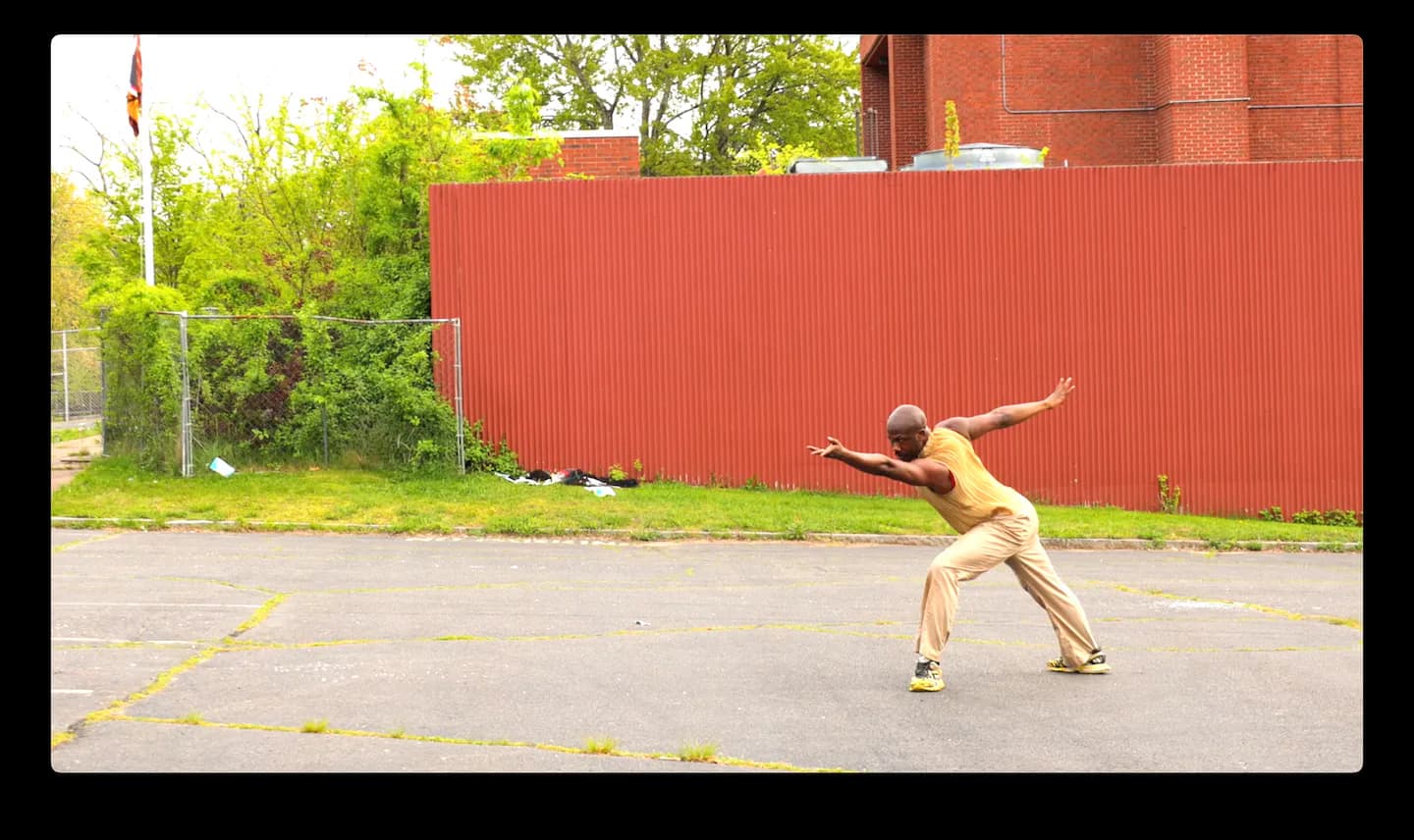 Photograph of a person in nude tones performing an inverted yoga or acrobatic pose on a concrete surface with a red corrugated metal wall and green foliage behind.