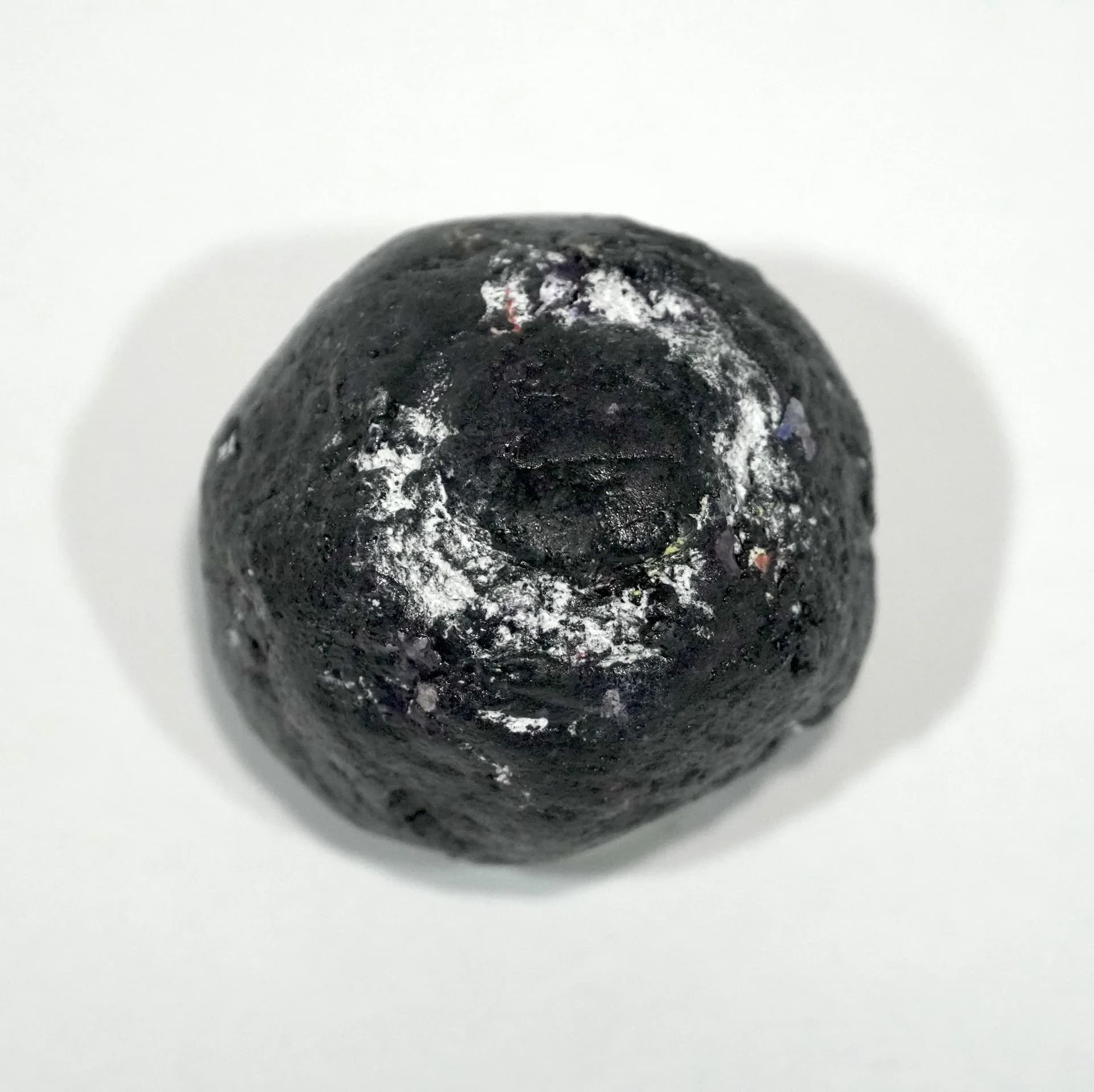 Overhead photograph of a dark charcoal-grey sphere with white powdery surface texture and a central circular depression, casting a soft shadow on white background.