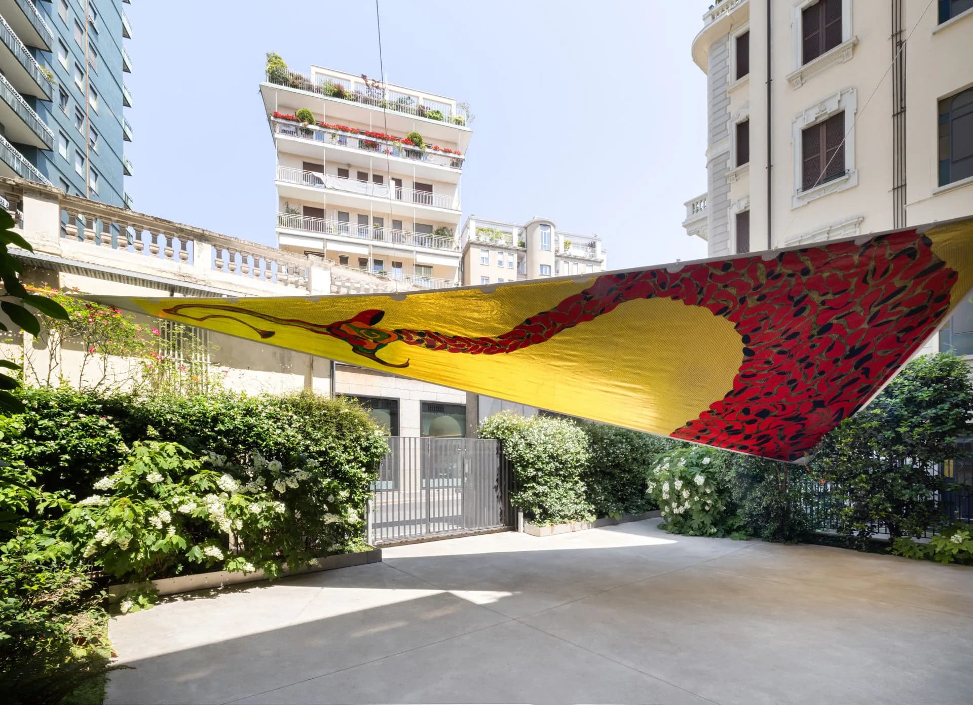 Outdoor sculptural installation with draped yellow and red fabric spanning between architectural elements, surrounded by green foliage in an urban courtyard.