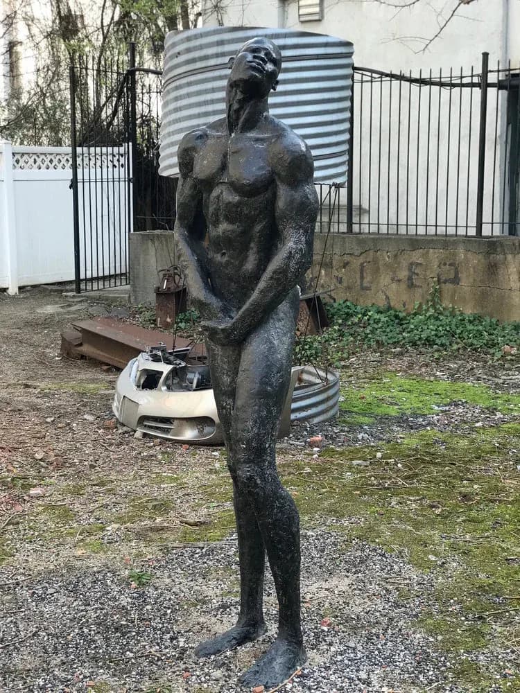 Bronze figurative sculpture of a standing female torso in dark patina, displayed in Kenkeleba House courtyard with corrugated metal backdrop and industrial objects.