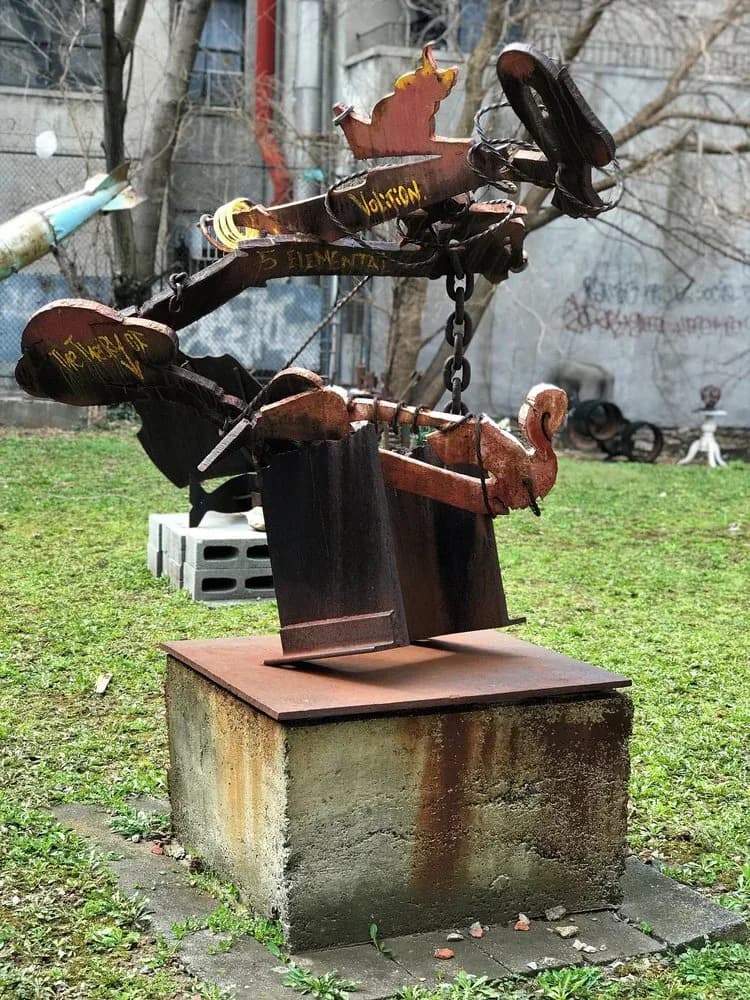 Rust-colored metal sculptural assemblage featuring organic and mechanical forms mounted on a concrete base in Kenkeleba House courtyard.
