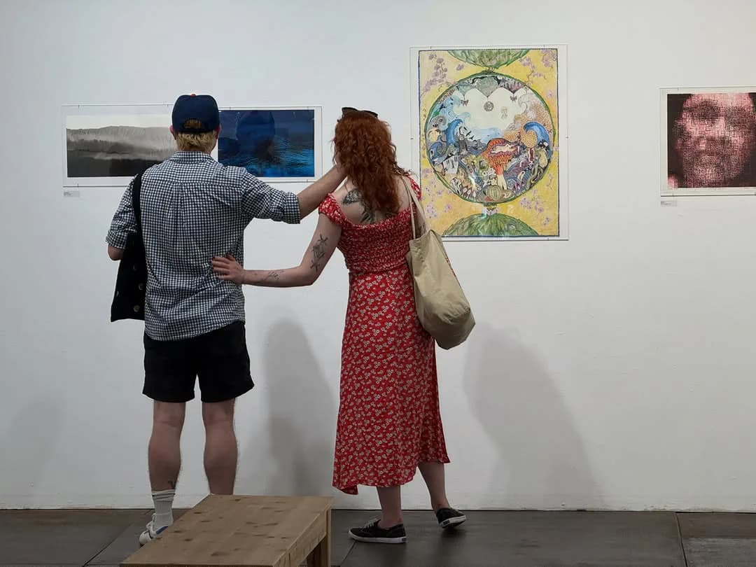 Two visitors in gallery space examining artworks on white walls, including a colorful circular illustration and dark paintings.