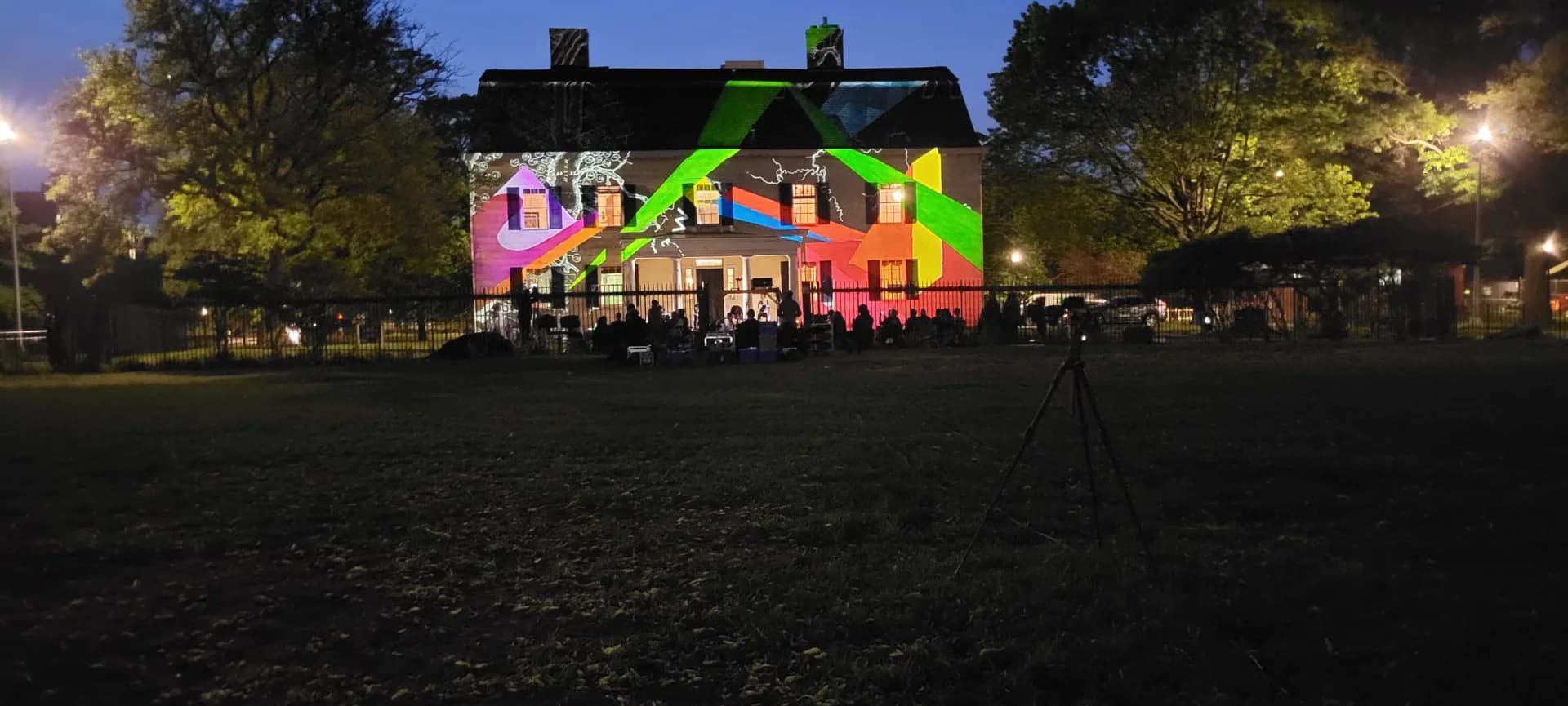 Colorful neon-bright projected light installation on a dark colonial building at dusk, with geometric shapes in pink, green, and blue.