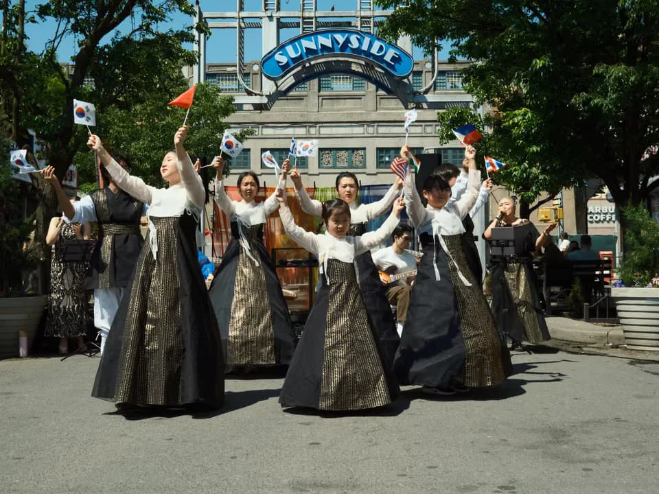 Performance art installation: figures in dark traditional robes with raised arms holding rainbow flags, standing beneath a Sunnyside archway.
