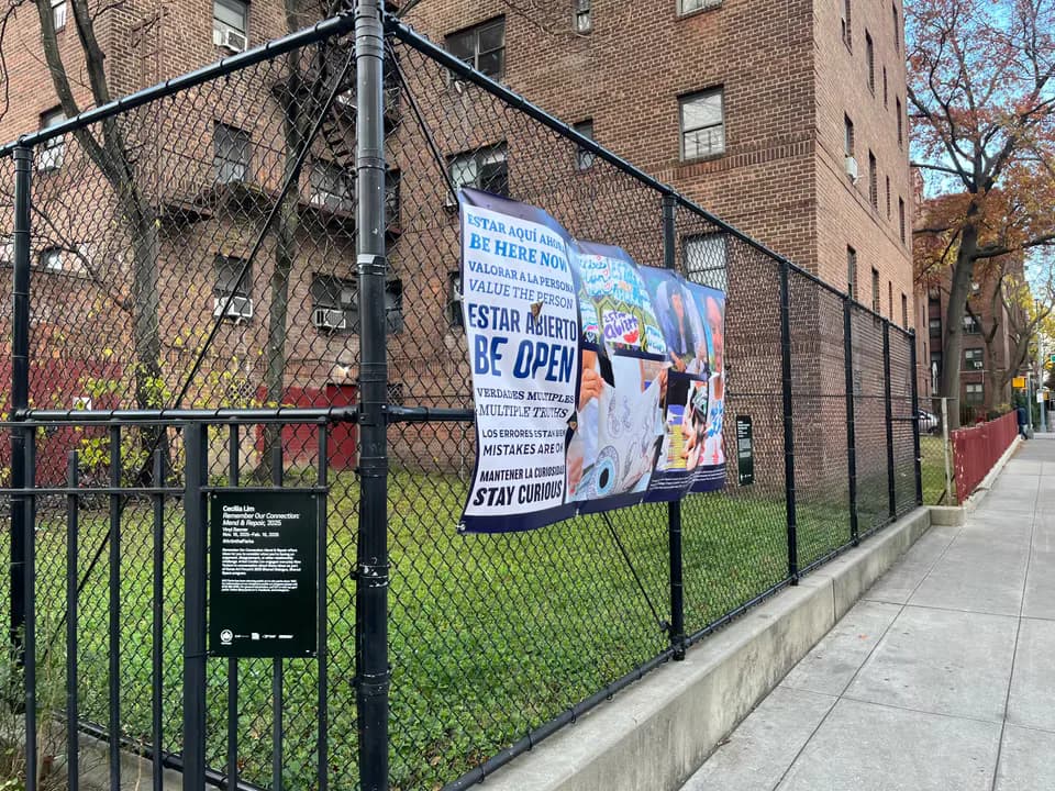 Black metal fence surrounding an outdoor community space with a blue and white banner announcing a reopening; brick buildings visible behind.