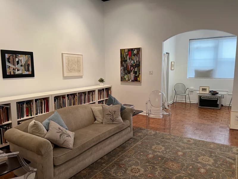 Living-room style gallery interior with a sofa, bookshelves, artwork on white walls, and a view into an adjoining room with parquet flooring
