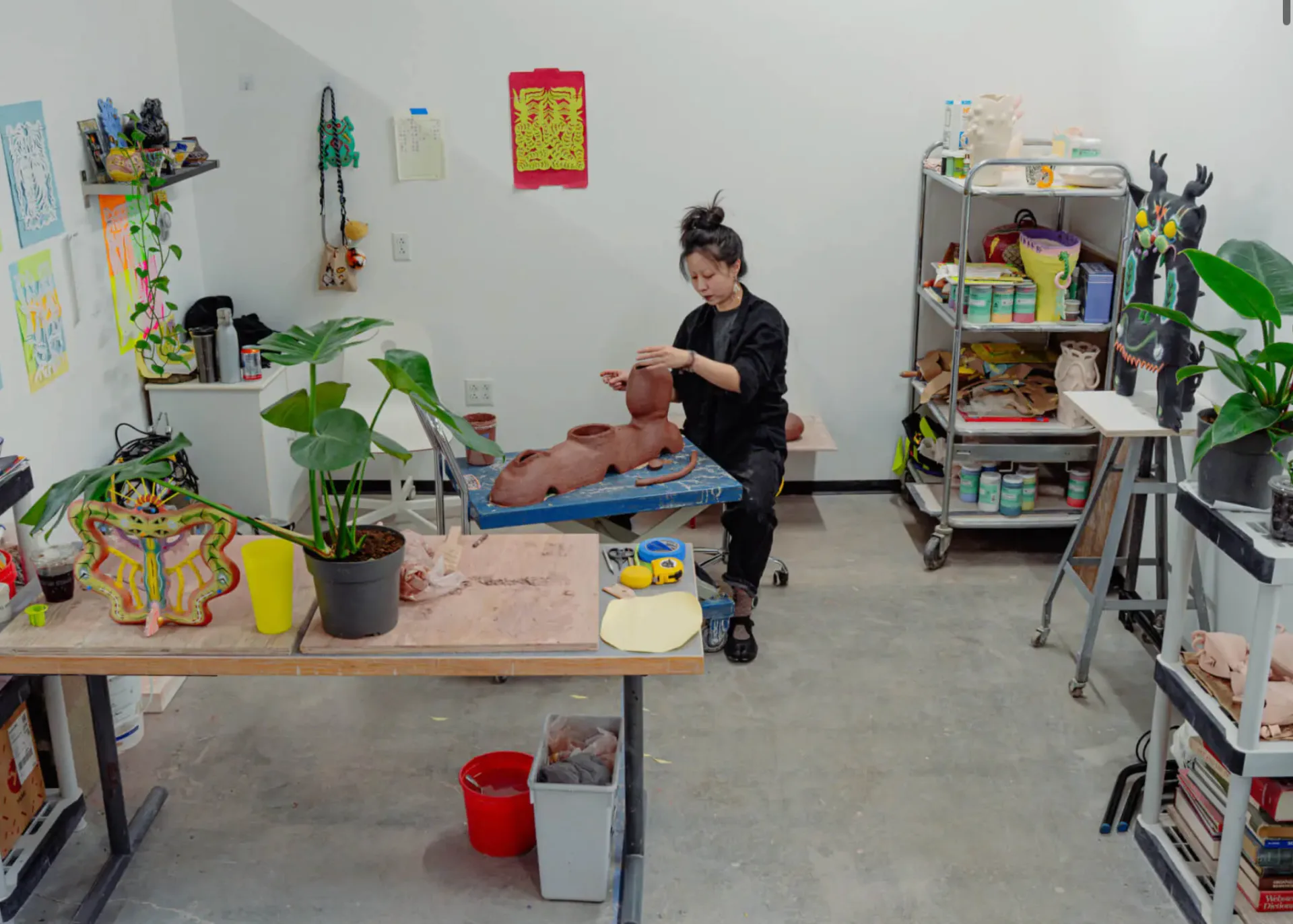 Artist working in a studio space surrounded by colorful ceramic vessels, potted plants, shelving units, and wall-mounted artwork.