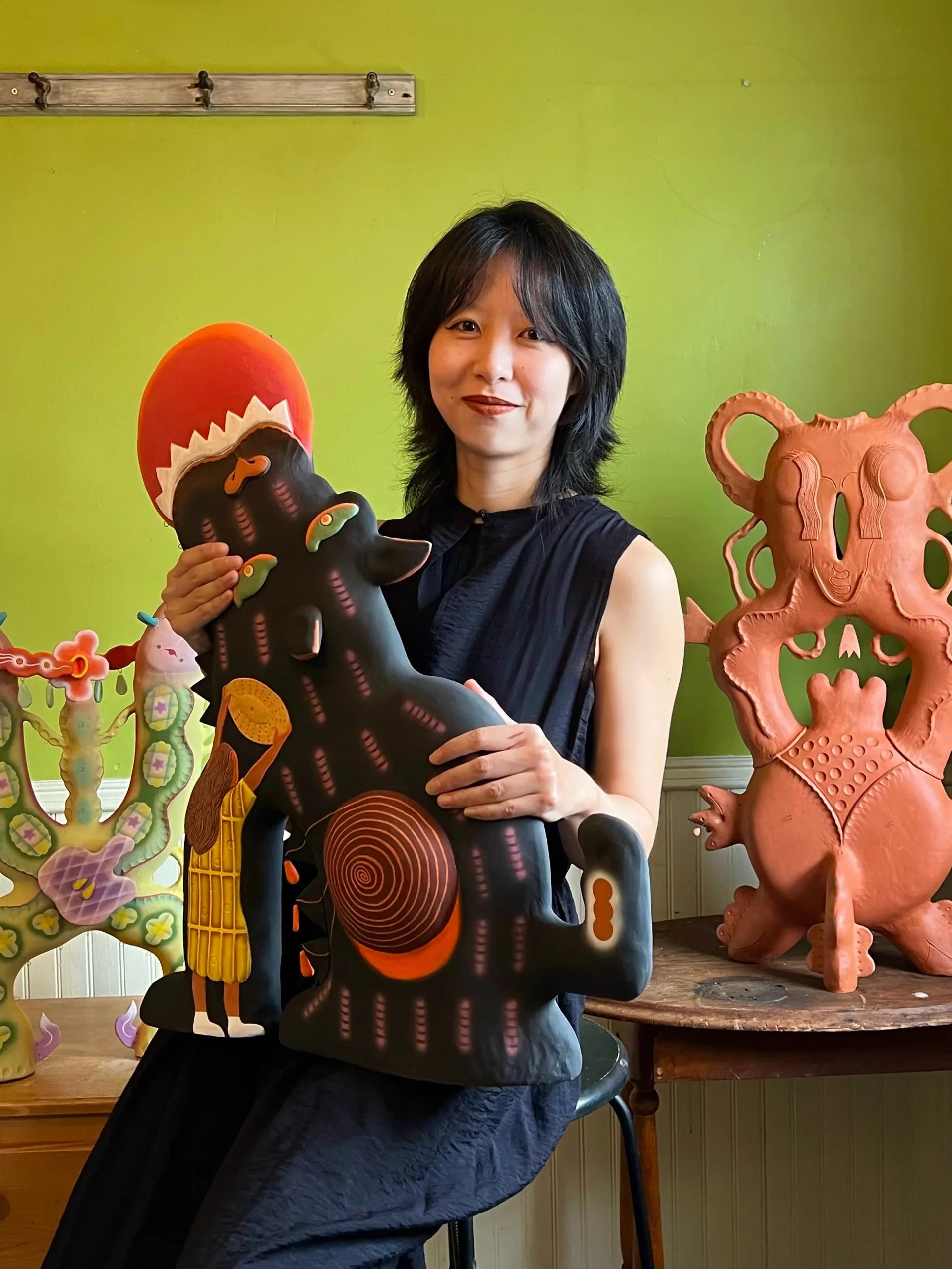Artist Kristen Lorello holding a large colorful ceramic sculptural piece, standing against a bright yellow-green wall with additional ceramic works visible.