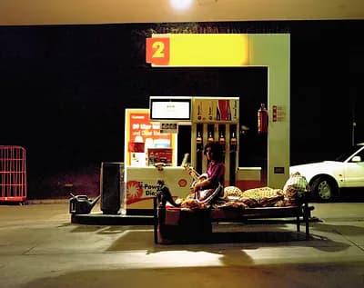 Installation artwork featuring a gas station storefront model with bright yellow-green signage and a white car, lit against dark surroundings.
