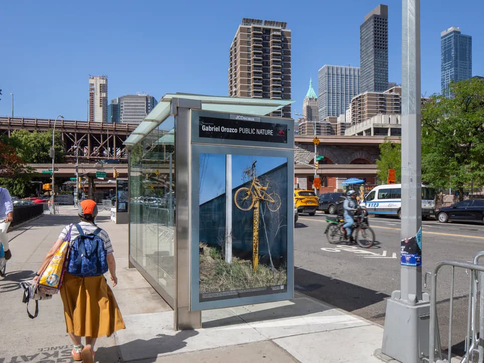 Street-level public art installation featuring a glass shelter with bicycle artwork, urban Chicago street with pedestrians and skyline in background.